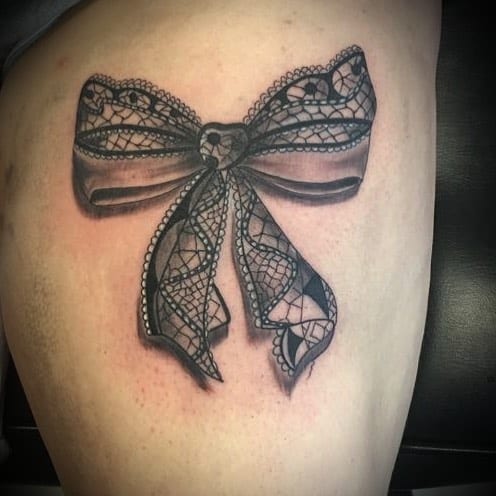 Black and grey thigh tattoo of a delicate lace bow with intricate patterns and shading, creating a soft, elegant, and realistic look.