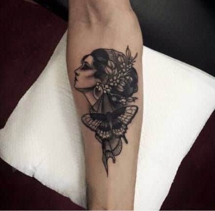 Black and grey tattoo of a gypsy woman in profile with floral hair ornaments and a detailed moth below, placed on the forearm.
