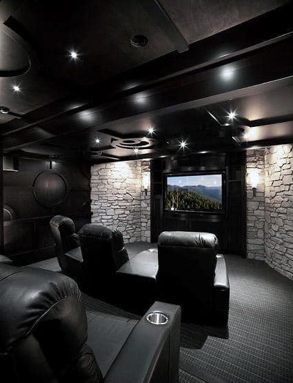 A cozy home theater with black leather recliners, stone walls, and a large screen displaying a scenic image
