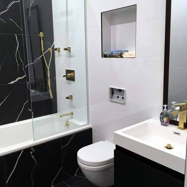 Modern bathroom with black marble tiles, gold fixtures, glass shower, and white vanity.