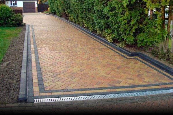 Brick-paved driveway with black concrete edging and a drainage system near landscaped hedges.