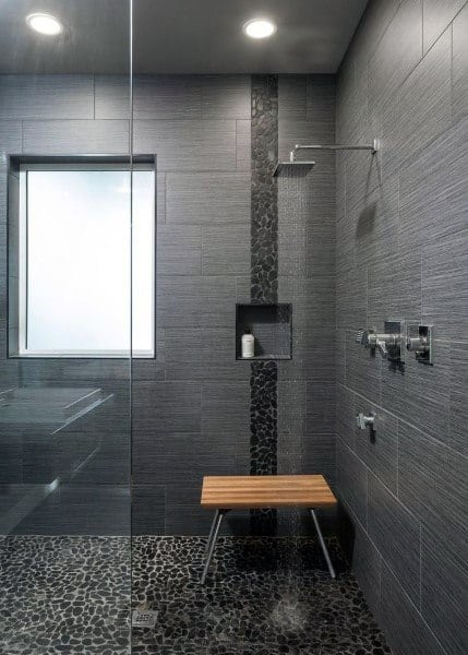 Modern shower with dark gray tiles, a wooden bench, pebble floor, and a sleek recessed shelf for a soap bottle