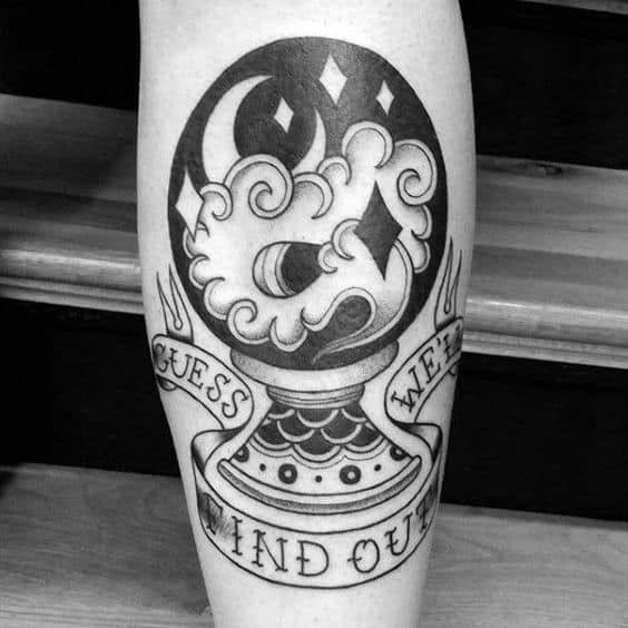 Black and grey tattoo of a crystal ball with swirling smoke, moon, and stars, featuring a banner that reads 'Guess We'll Find Out' on the leg.