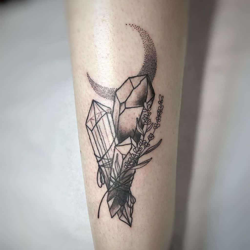 A tattoo on the forearm featuring geometric crystals wrapped with fine lines, lavender sprigs, and a crescent moon, all rendered in black ink with dotwork shading.