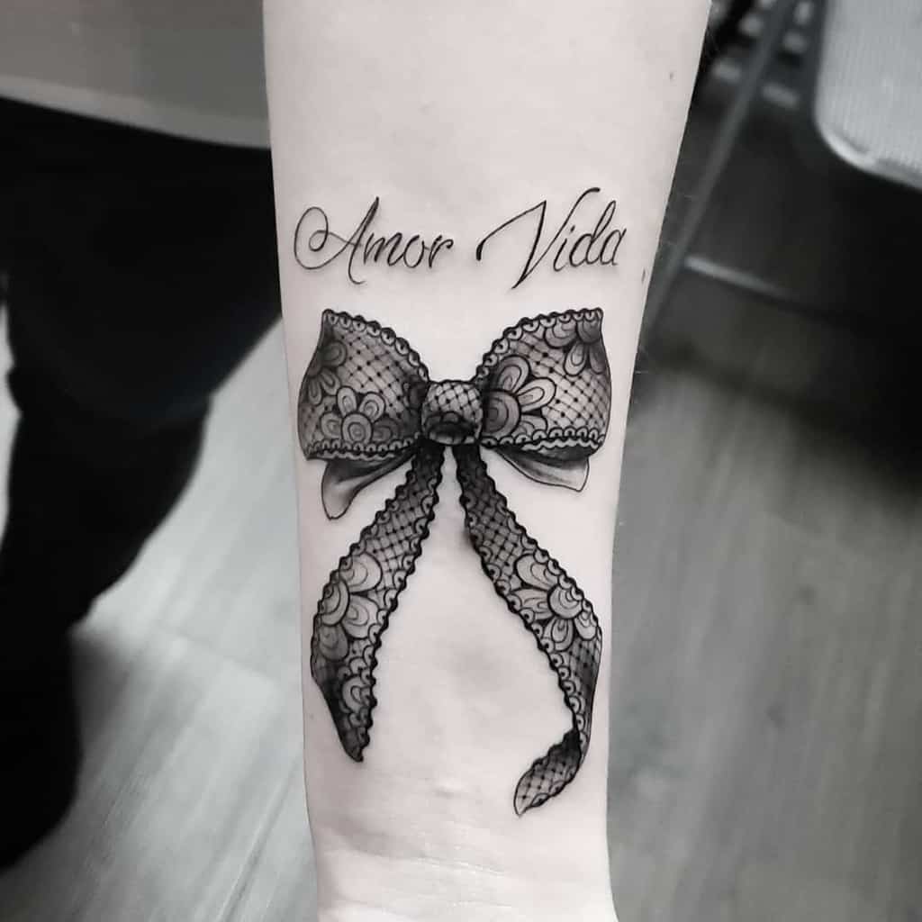 Black lace bow tattoo on the forearm with floral patterns, delicate shading, and the phrase 'Amor Vida' scripted above it.