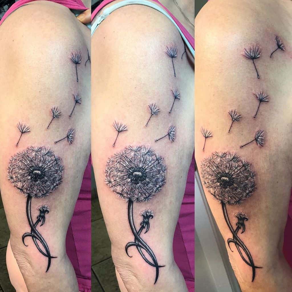 A black and grey dandelion tattoo on the upper arm with seeds floating away, featuring intricate linework and shading details.