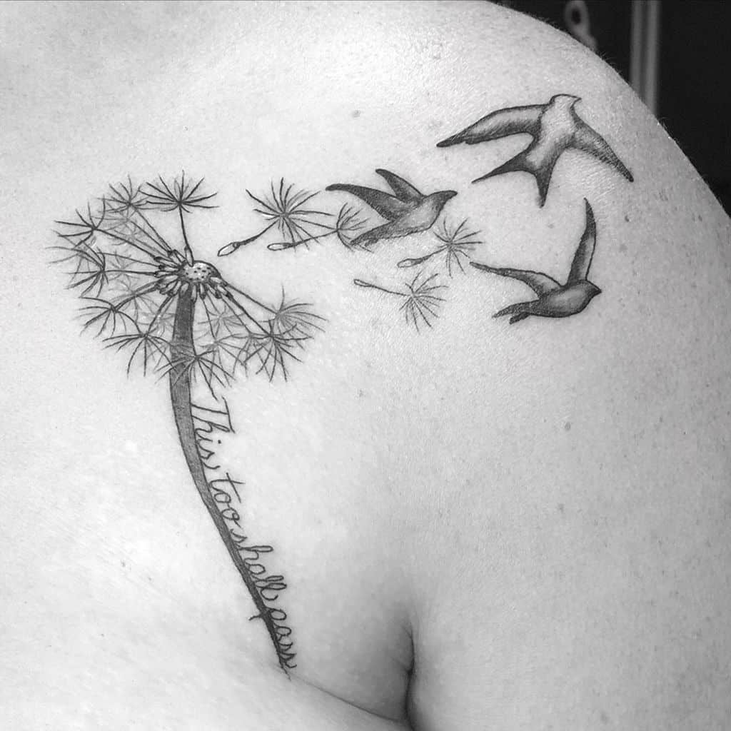 A black and white tattoo of a dandelion, seeds blowing away, with birds flying above and the words "This too shall pass" beneath.
