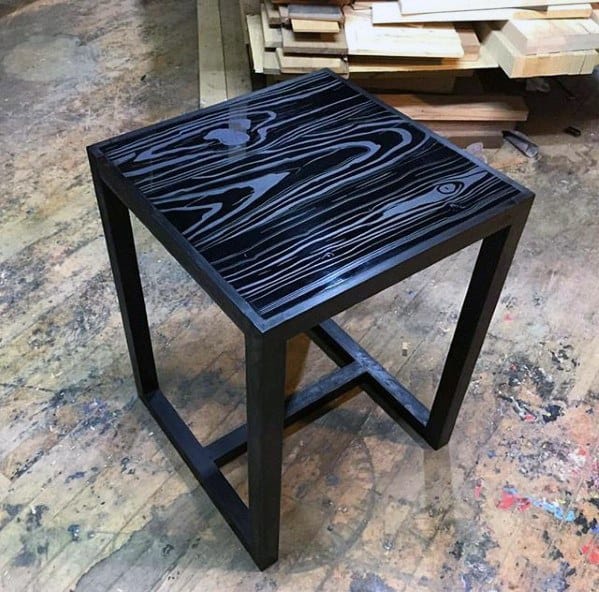 Square black wooden side table with a textured, wood-grain top, set on a concrete floor