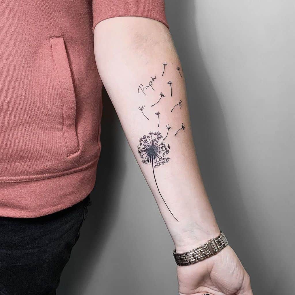A dandelion tattoo on the forearm with floating seeds and the word "Papa" written in a cursive font above the flower.