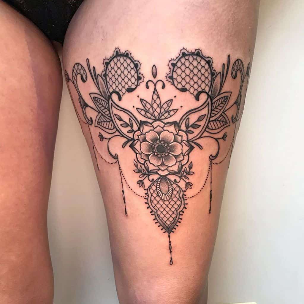 Elegant black ink thigh tattoo featuring intricate lace patterns, floral elements, ornamental filigree, and delicate beaded details.