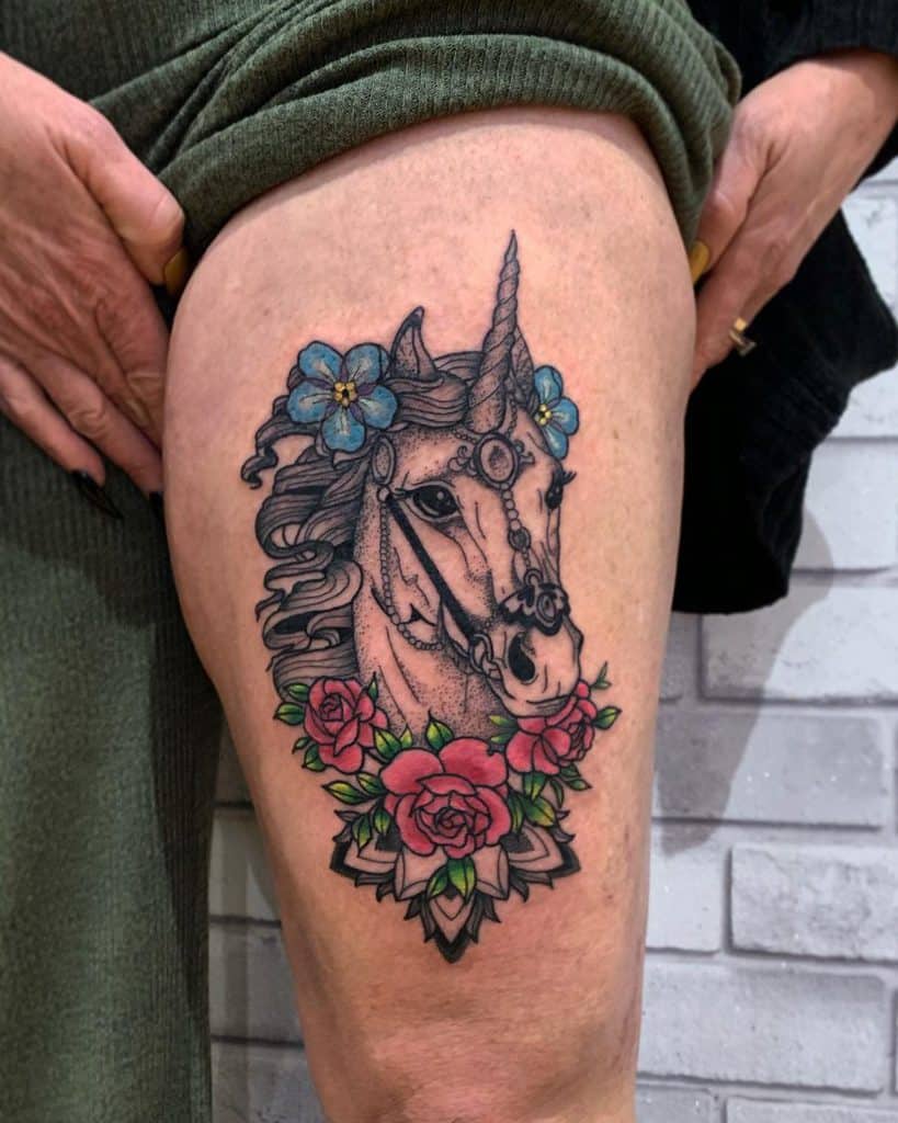 Colorful thigh tattoo of a unicorn with intricate jewelry, blue and red flowers, and bold black outlines, blending fantasy with a classic tattoo style.