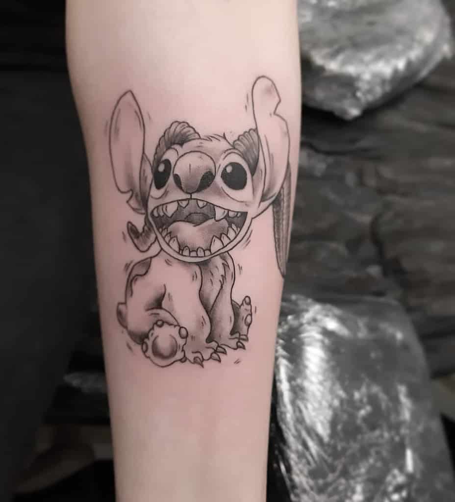 Black and white tattoo of Stitch from Lilo & Stitch with a big grin and playful expression, sitting on the arm.
