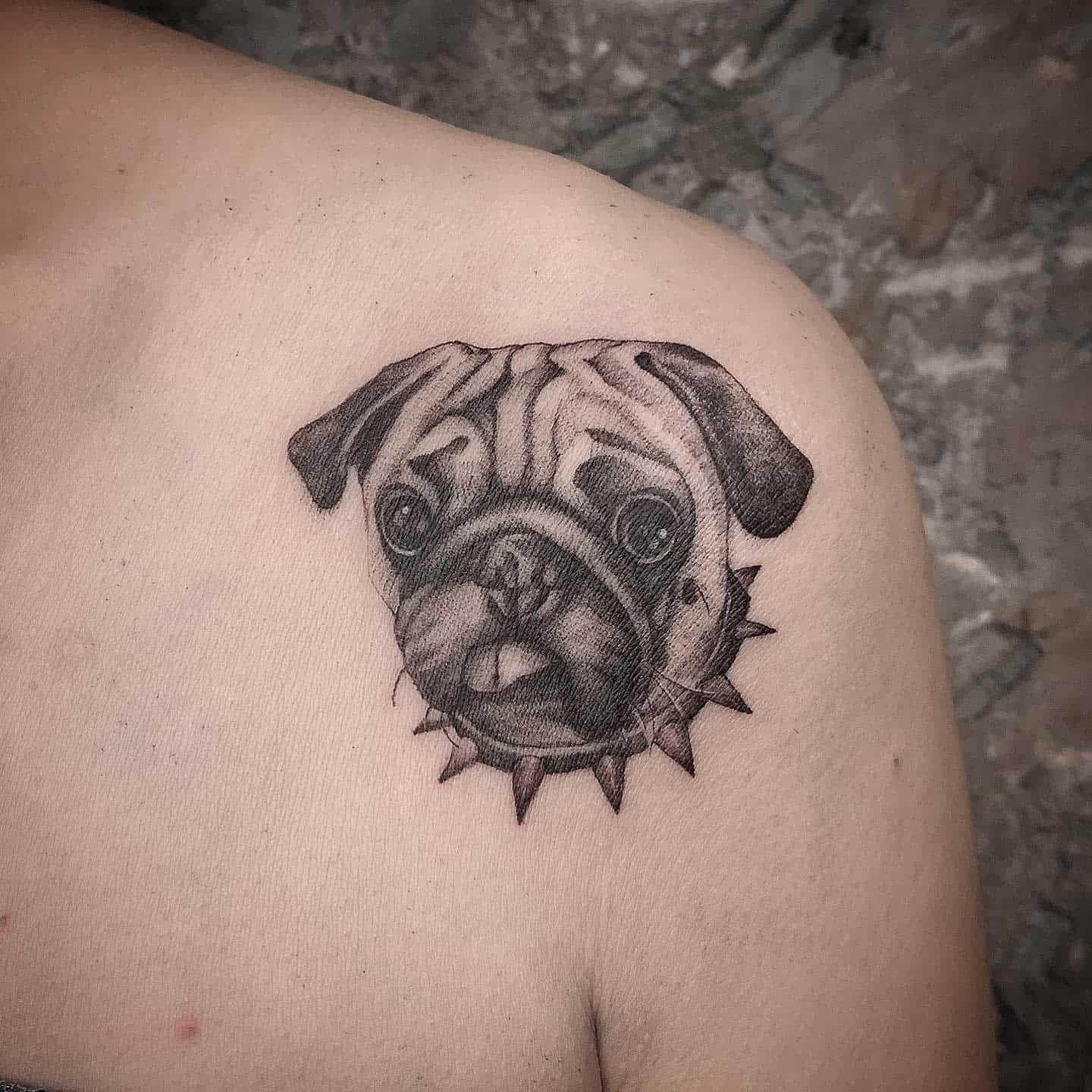 Black and grey tattoo of a pug's face, showing the tongue with a spiked collar 