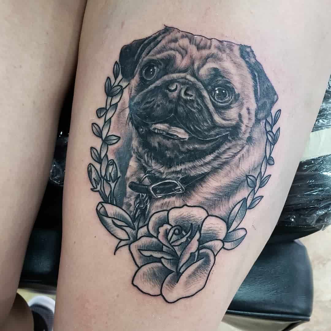 A detailed black-and-white tattoo of a smiling pug surrounded by floral elements, including a rose and leaves, with a collar