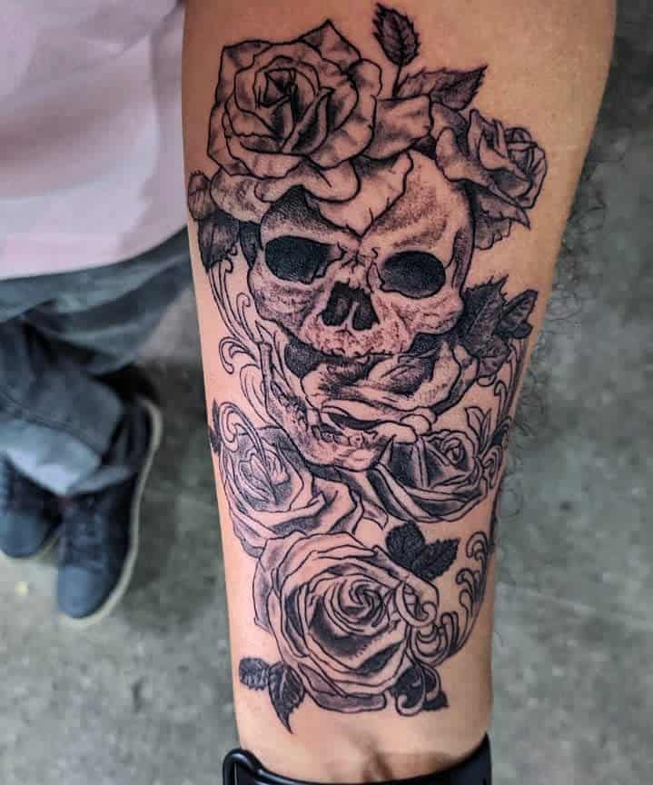 Intricate black and grey tattoo of a cracked skull surrounded by detailed roses and swirling patterns, covering the forearm.