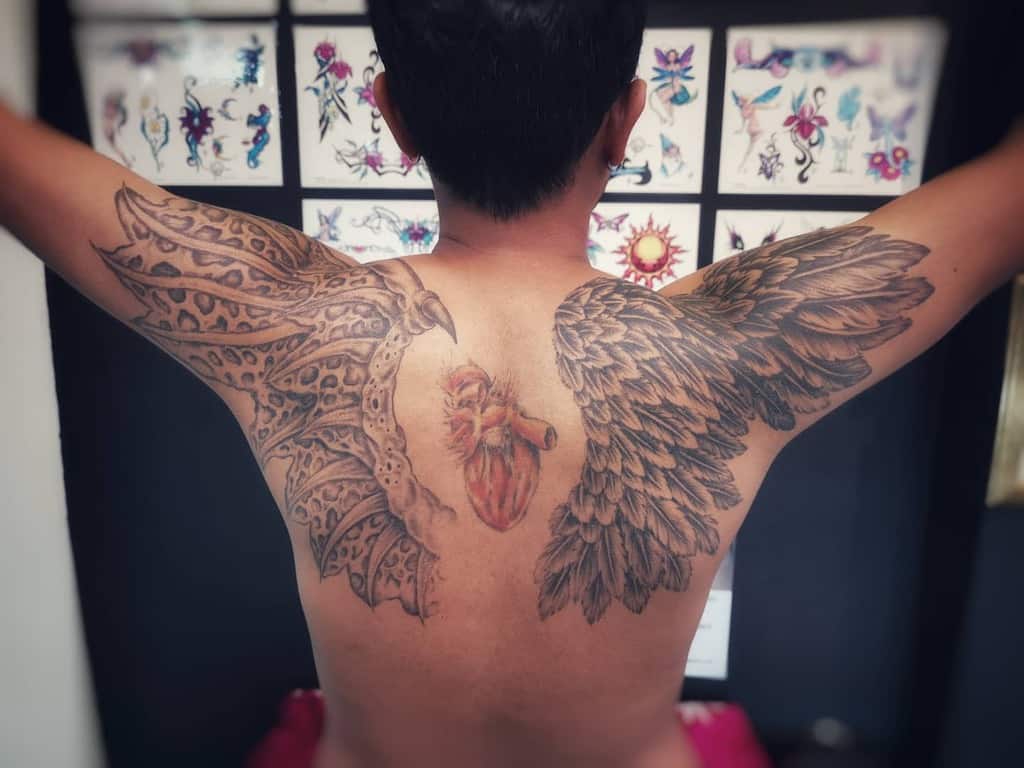 Unique tattoo featuring a detailed angel wing on one side and a demonic, reptilian wing on the other, with a realistic heart in the center