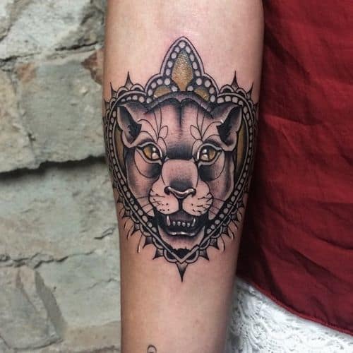 Forearm tattoo of a fierce lioness framed by an ornate mandala-style design, featuring bold black ink and gold accents.
