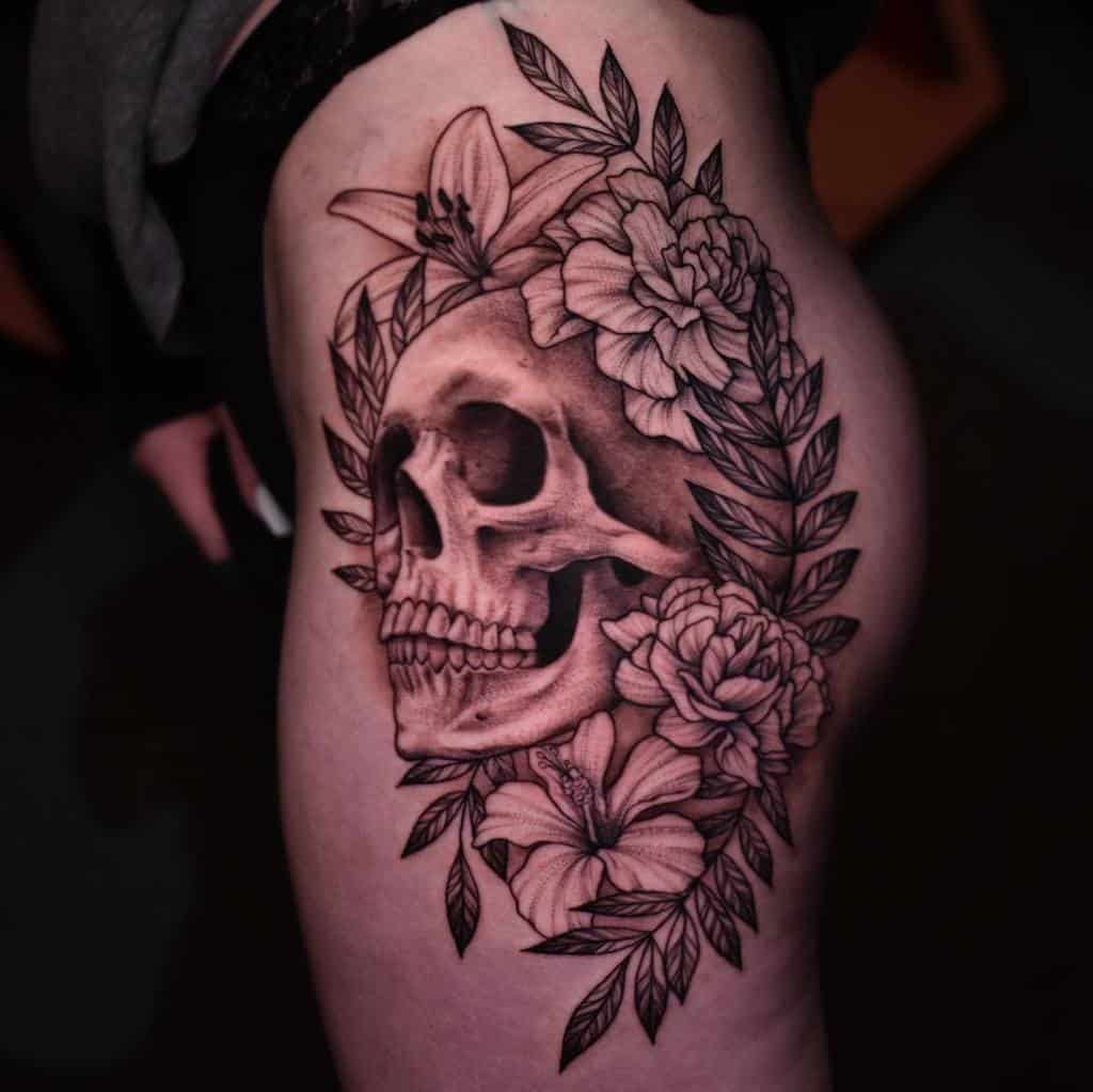 Black and grey thigh tattoo featuring a detailed skull surrounded by blooming flowers and leaves, creating a striking contrast
