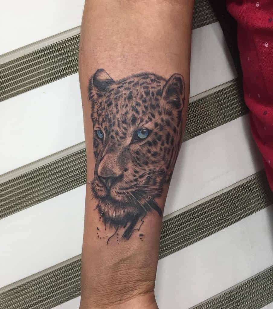 A realistic tattoo of a jaguar's face with striking blue eyes, positioned on the forearm with detailed shading.