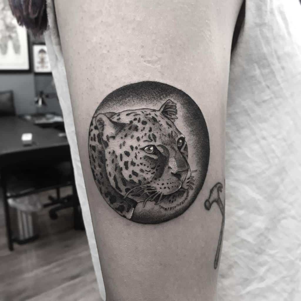 A striking black and grey tattoo of a jaguar's face within a circular frame, placed on the upper arm with smooth shading and realistic details.