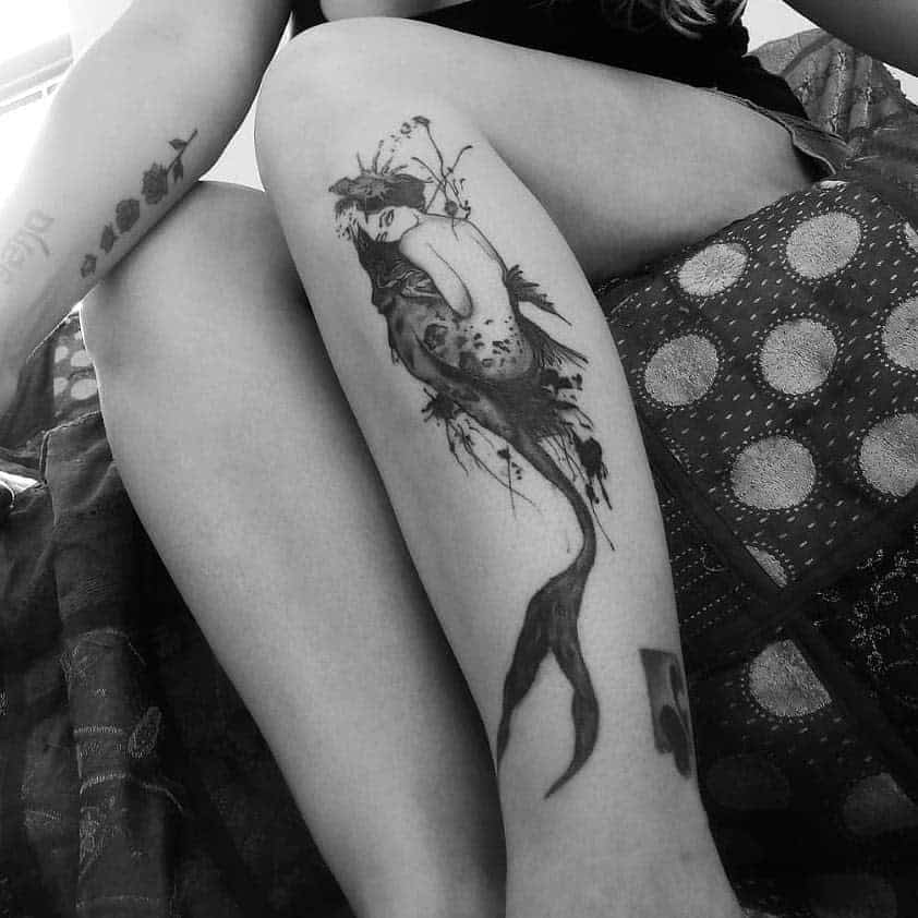 Black and grey tattoo of a mystical mermaid with abstract watercolor-style shading, delicate facial details, and a flowing, ethereal tail.
