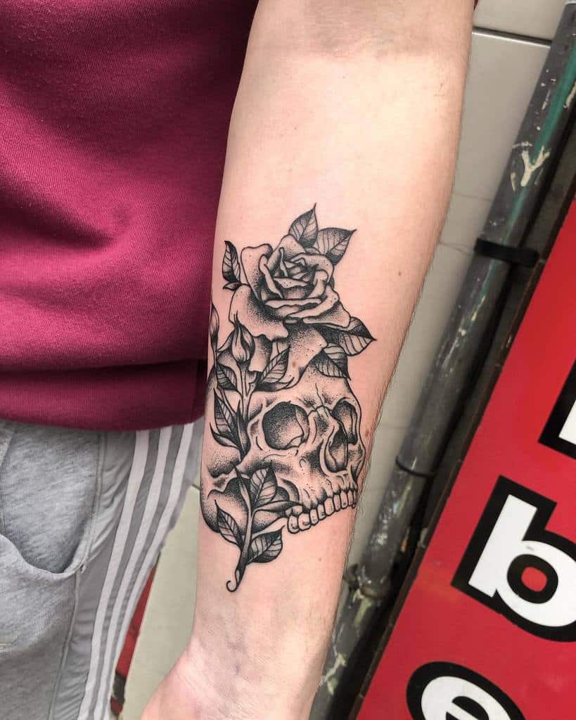 Black and grey forearm tattoo of a skull intertwined with roses and leaves, featuring fine linework and detailed shading.