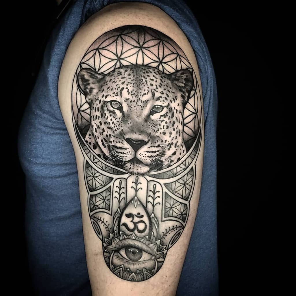 A detailed black and gray tattoo featuring a jaguar's face framed by geometric patterns, with symbols like the Om, an eye, and a lotus, creating a spiritual and mystical design.