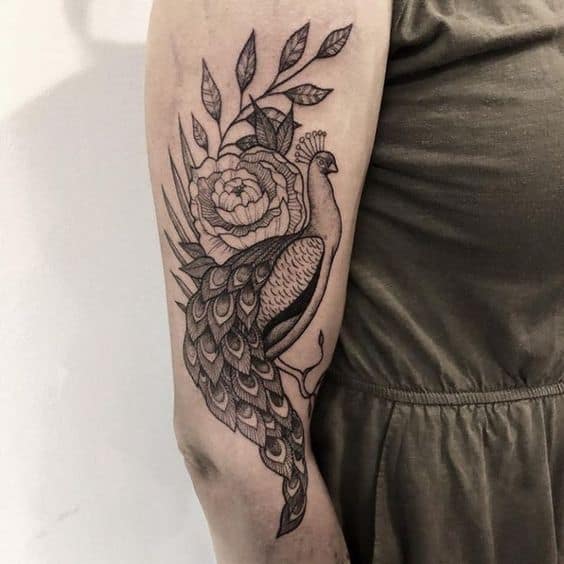 Black ink peacock tattoo on the upper arm, featuring intricate feather details, floral elements, and fine line shading for depth.