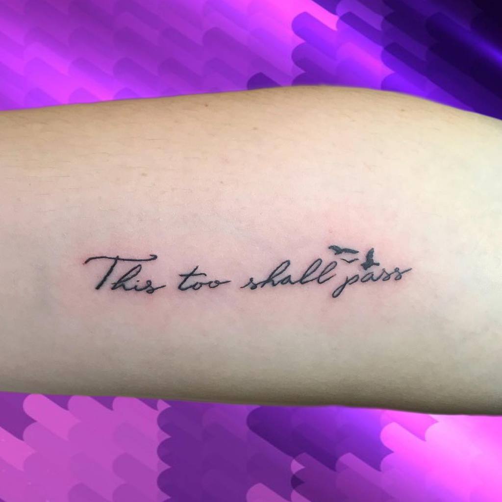 A "This Too Shall Pass" tattoo in cursive with a small bird design, on a person's forearm.