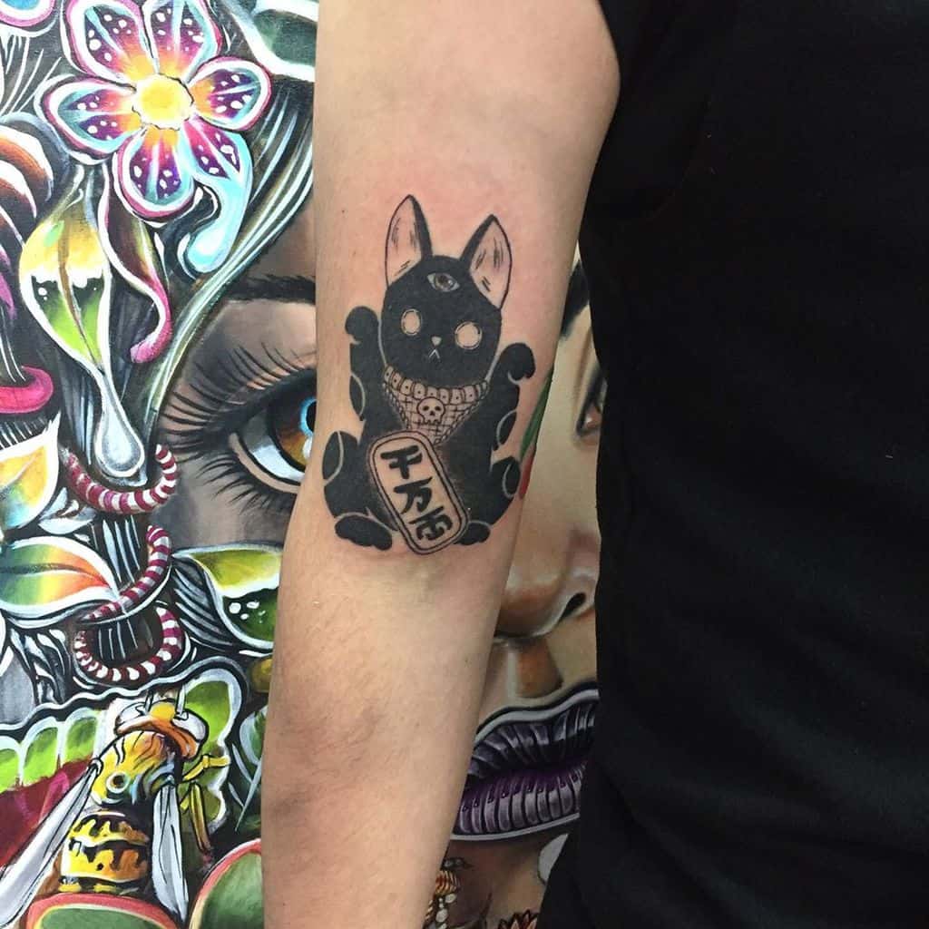 Tattoo of a dark Maneki-Neko cat with a third eye, skull-like grin, and Japanese kanji on its coin, inked on the upper arm.