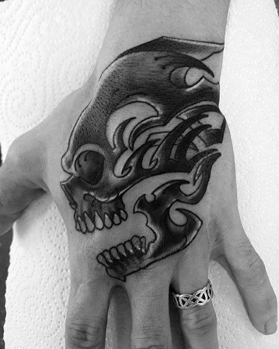 Bold tribal skull tattoo on the back of a hand, featuring sharp black patterns and stylized flames integrated into the skull.