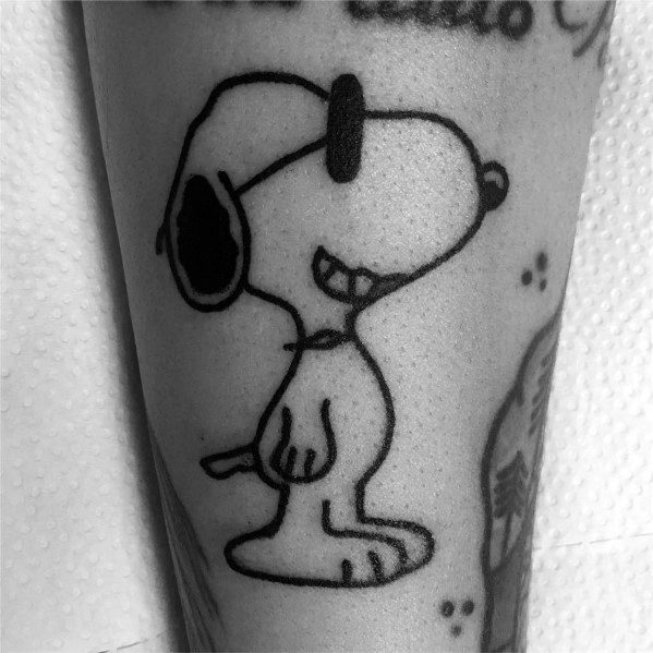 Black and white tattoo of Snoopy standing with a big grin and wearing sunglasses, placed on a person's arm.
