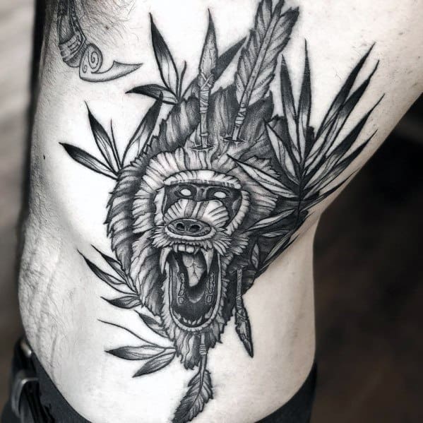 Black and grey baboon tattoo with tribal-inspired elements, bold shading, and fierce expression, featuring feathers and intricate details on the ribs.