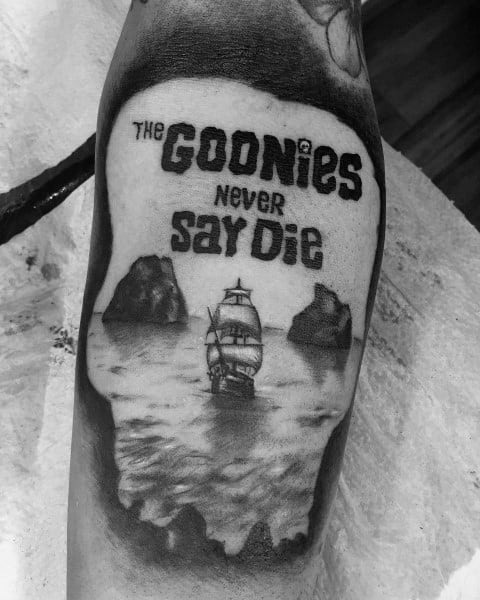 Black and gray tattoo of The Goonies featuring a skull-shaped design with a pirate ship, rocky islands, and the phrase "Goonies Never Say Die".