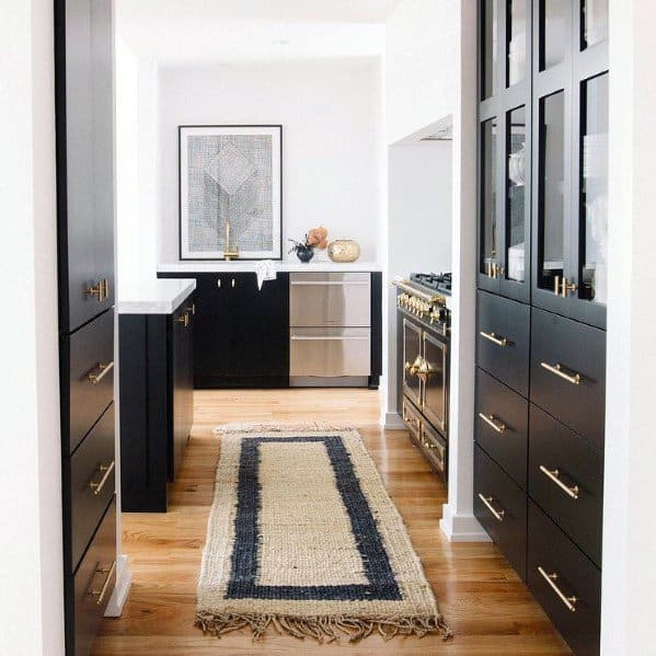 Black kitchen cabinets with gold hardware, wood floors, and a woven runner rug.