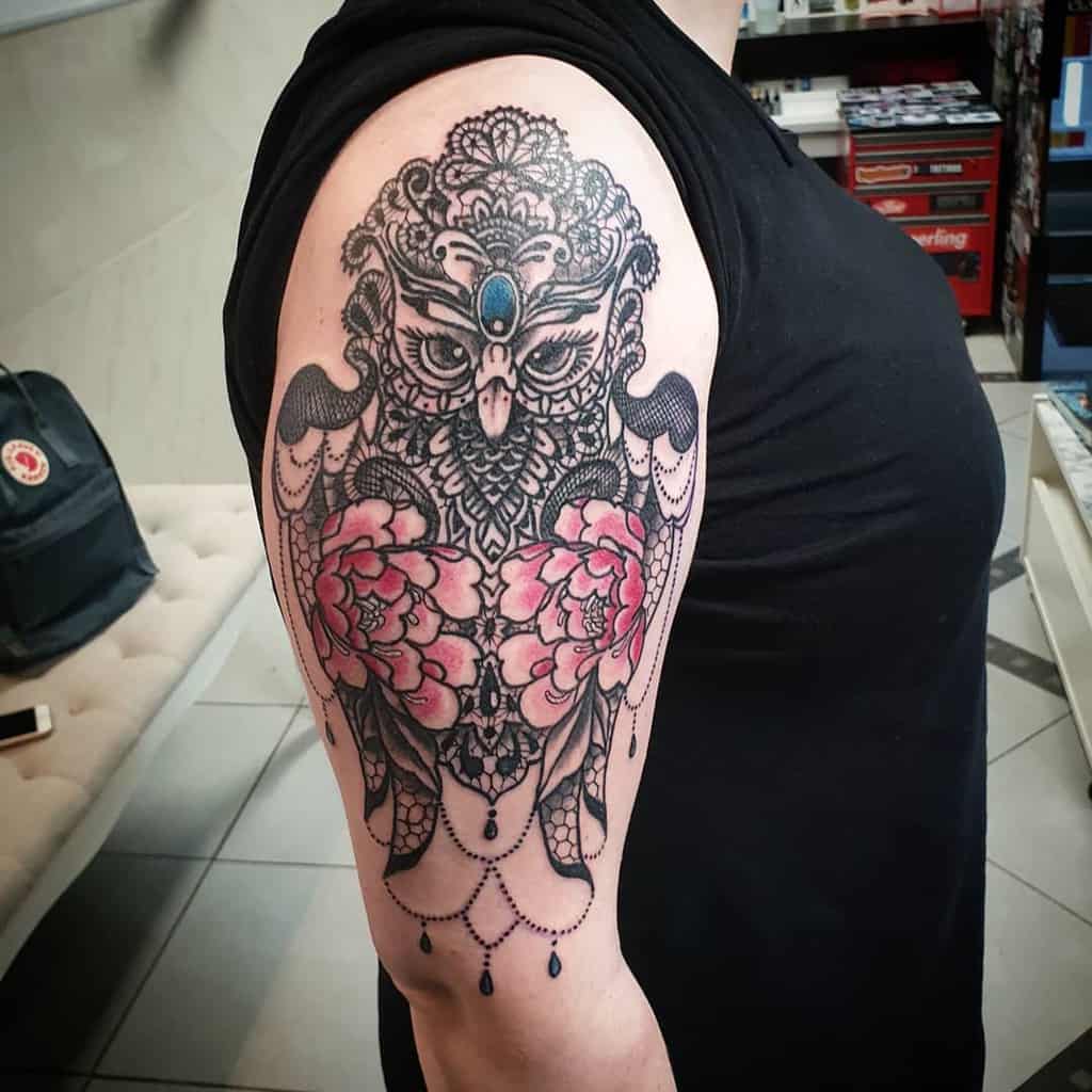 Ornate upper arm tattoo featuring a symmetrical owl design with intricate lace details, pink peonies, a gemstone centerpiece, and beaded chains.