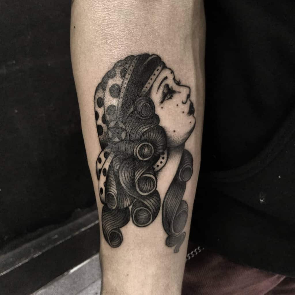 Black and grey tattoo of a gypsy woman with voluminous curled hair, polka dot headscarf, and delicate shading on the forearm.