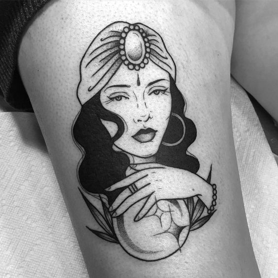 Black and grey tattoo of a mystical gypsy woman with a jeweled headscarf, hoop earrings, and a crystal ball on the thigh.