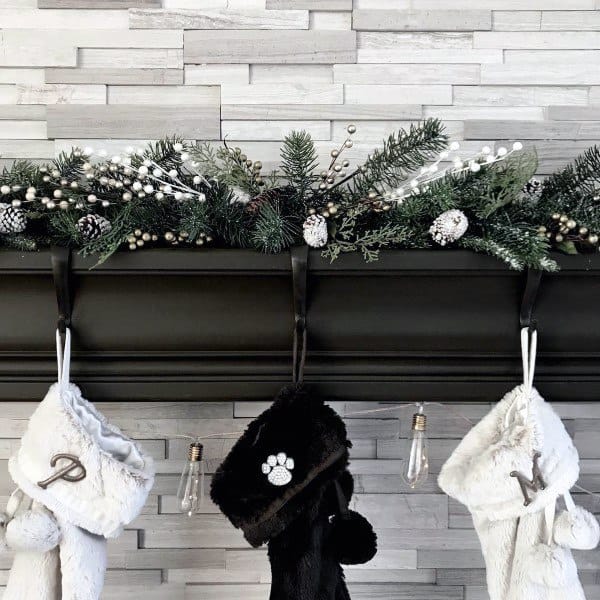 Black fireplace mantel with festive garland, hanging stockings, and a stone tile wall backdrop.