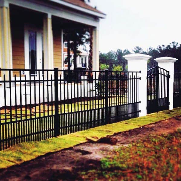 A sleek black metal fence with white posts encircles a home featuring white pillars and a mustard exterior