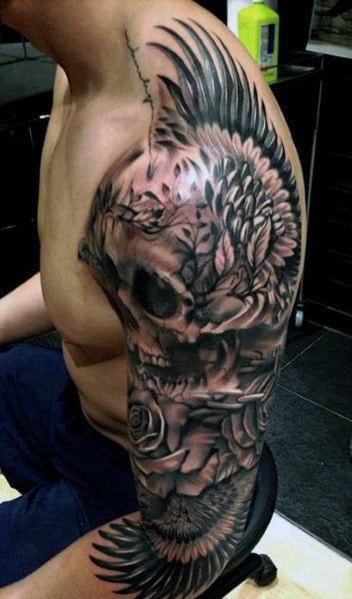 Full-sleeve tattoo featuring a skull, roses, and winged elements, with dark shading and intricate detailing for a bold design