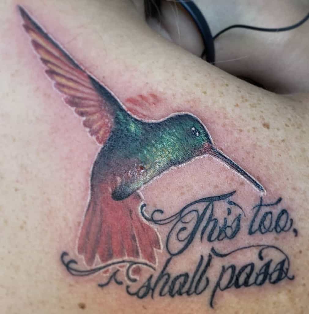 A "This Too Shall Pass" tattoo in cursive with a colorful hummingbird design, on a person's upper back.