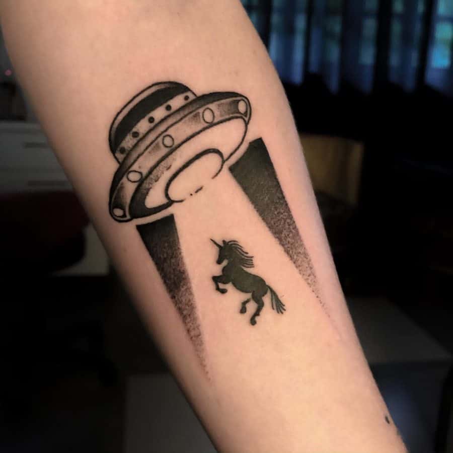Black ink tattoo of a UFO beaming up a unicorn, blending sci-fi and fantasy in a surreal design on the forearm.