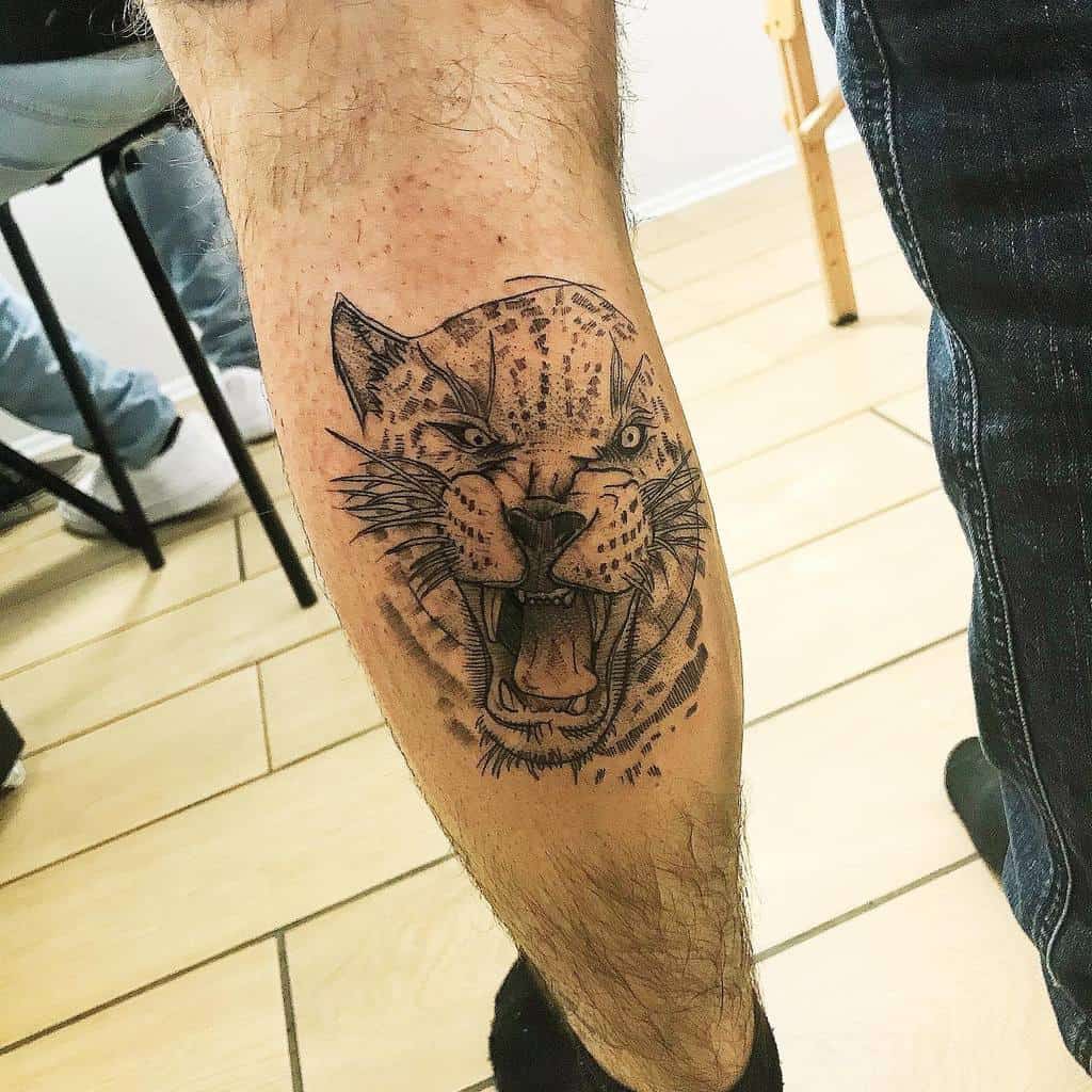A detailed black ink tattoo of a snarling jaguar, with intense facial features and sharp lines, placed on the lower leg for a bold and fierce design.