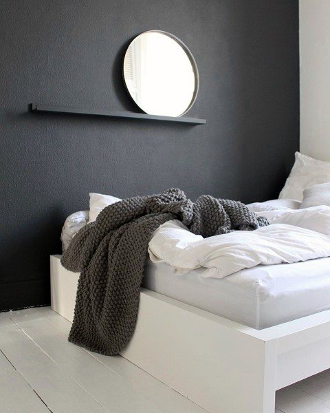 Minimalist black bedroom with white bedding, knit throw, and round mirror on textured wall.