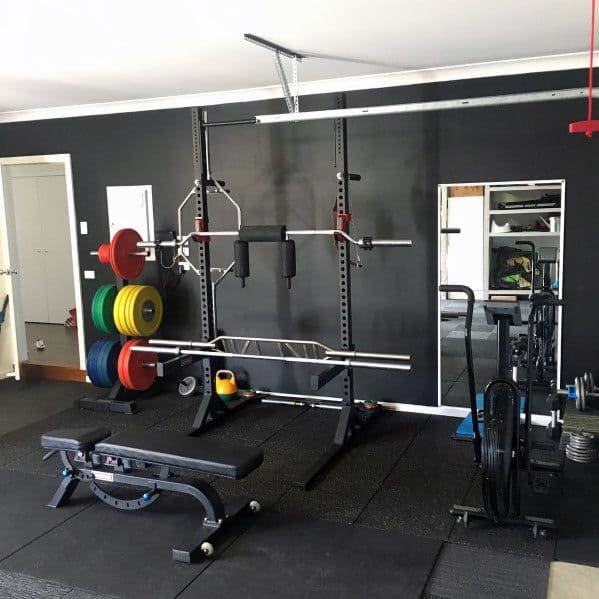 Garage gym with a black squat rack, weight plates, adjustable bench, and air bike on rubber flooring.