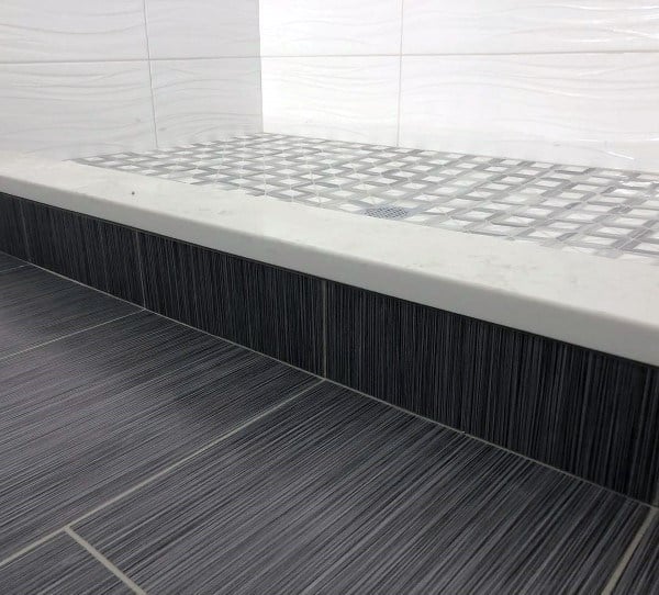 Black striped floor tiles with a white marble shower curb and textured white wall tiles.