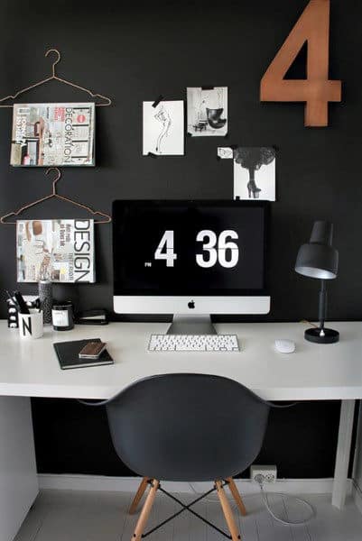 Minimalist workspace with a black wall, a desk with an iMac showing 4:36, magazines, art, and a lamp