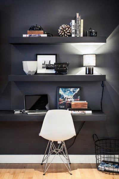 Minimalist desk with white chair, shelves holding books, a lamp, and decor items against a dark wall