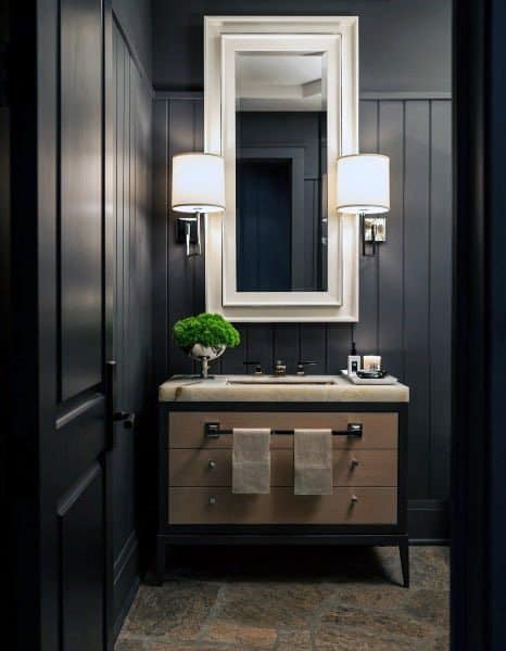 A modern half bath with dark walls, a large mirror, twin sconces, and a vanity with towels and a small plant